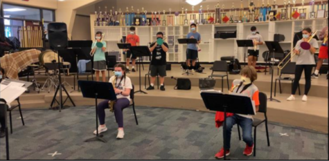 High school band students play their instruments in the band room. They are socially distanced with masks around their instruments.
